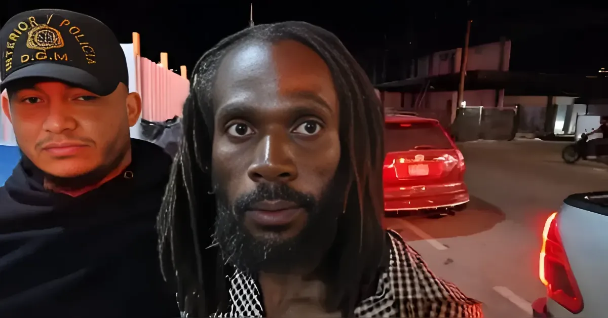 Two men posing at night on a street; foreground man has long dreadlocks and a beard, wearing a black-and-white checked shirt, while a second man in a cap and hoodie stands beside him.
