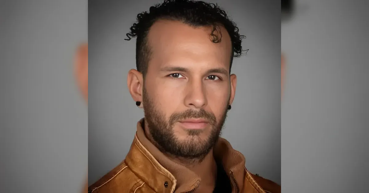 Portrait of a man with dark hair and a short beard, wearing a tan leather jacket, looking directly at the camera against a gray background.