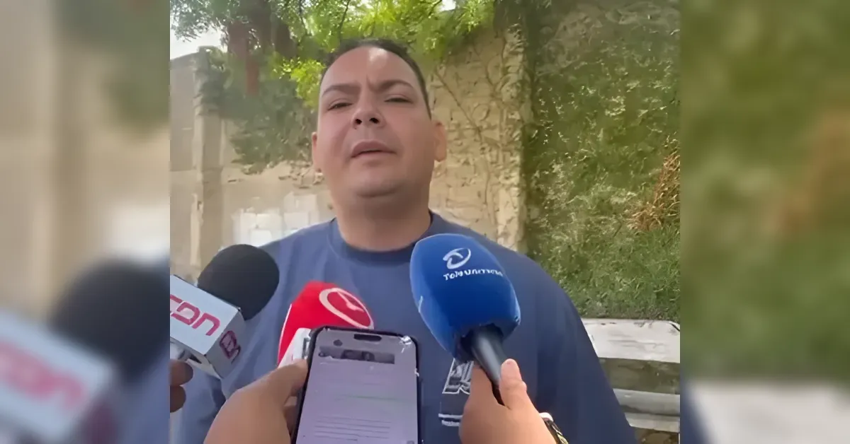 Man being interviewed by multiple reporters outdoors, microphones held close to him as cameras record the statement