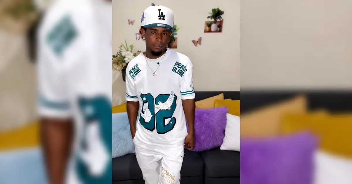 Young man in a white sports-inspired outfit stands in a living room, wearing an LA cap and a shirt with 'Peace Be Love' and the number 92, with yellow and purple cushions behind him.