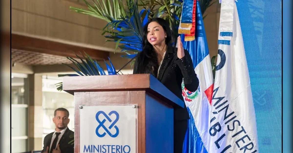 Speaker at a wooden podium delivering a speech, with a ministry logo on the front and Dominican flags in the background.