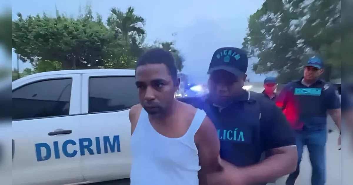 Officer in a dark vest escorts a man in a white tank top beside a police pickup marked DICRIM, blue emergency lights in the background, outdoors.