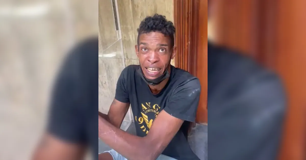 Young man with short curly hair in a black T-shirt, mask under his chin, seated indoors near a wooden door, looking up with an expression of concern.