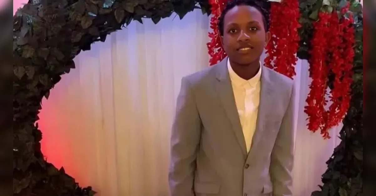 Person in a light gray suit and white shirt posing under a leafy arch with red hanging decorations at a formal event .