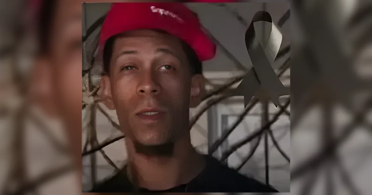 Portrait of a young man wearing a red cap, facing the camera with a blurred metal fence in the background and a gray ribbon overlay in the foreground.