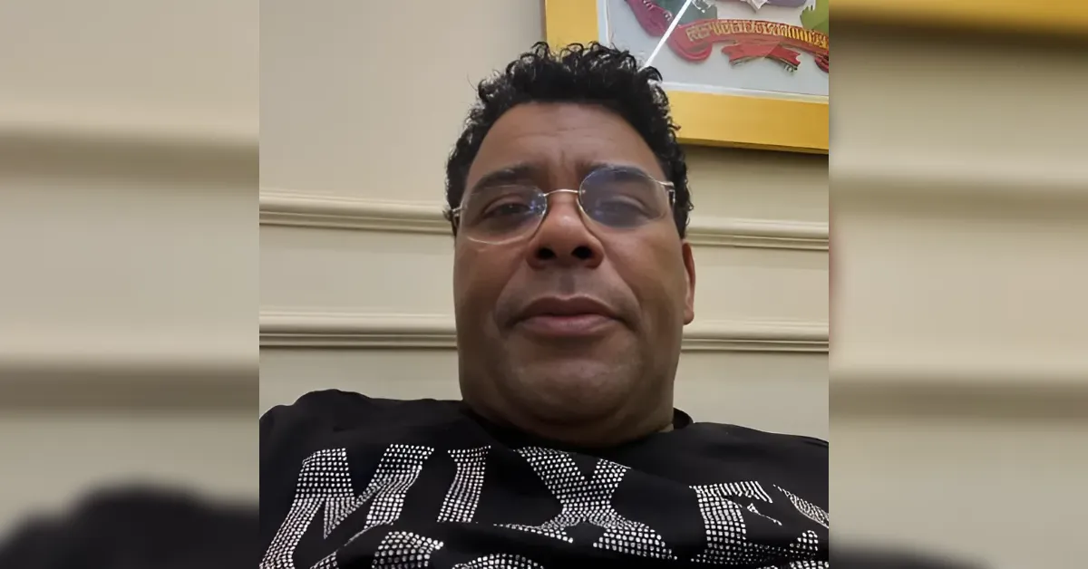 Close-up selfie of a man wearing round glasses, looking at the camera indoors with a framed artwork behind him.