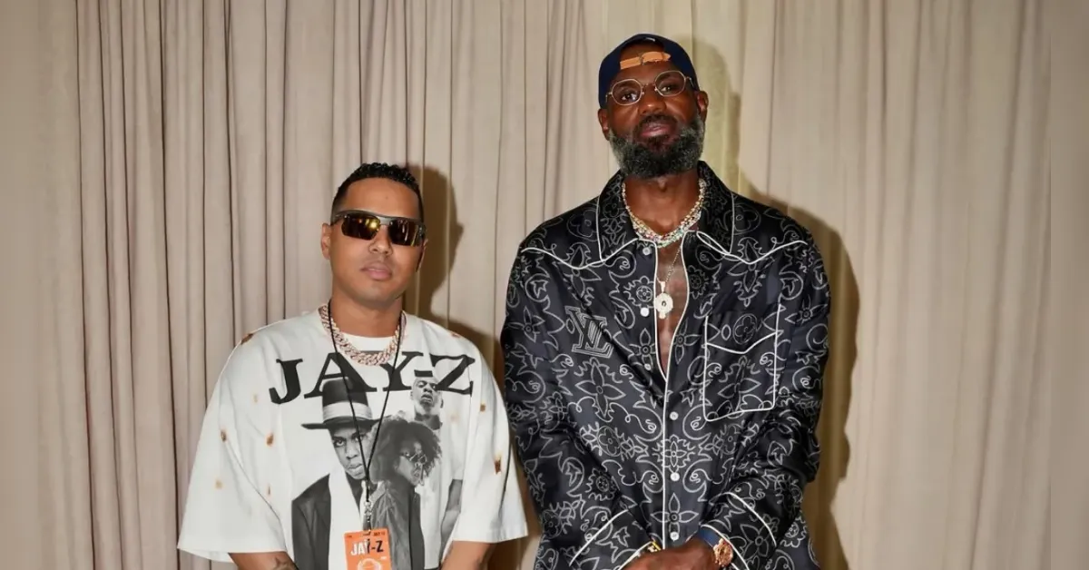 Two men posing side by side in front of beige curtains; left man wears a white graphic T‑shirt and sunglasses, right man in a patterned black shirt and cap with chains.