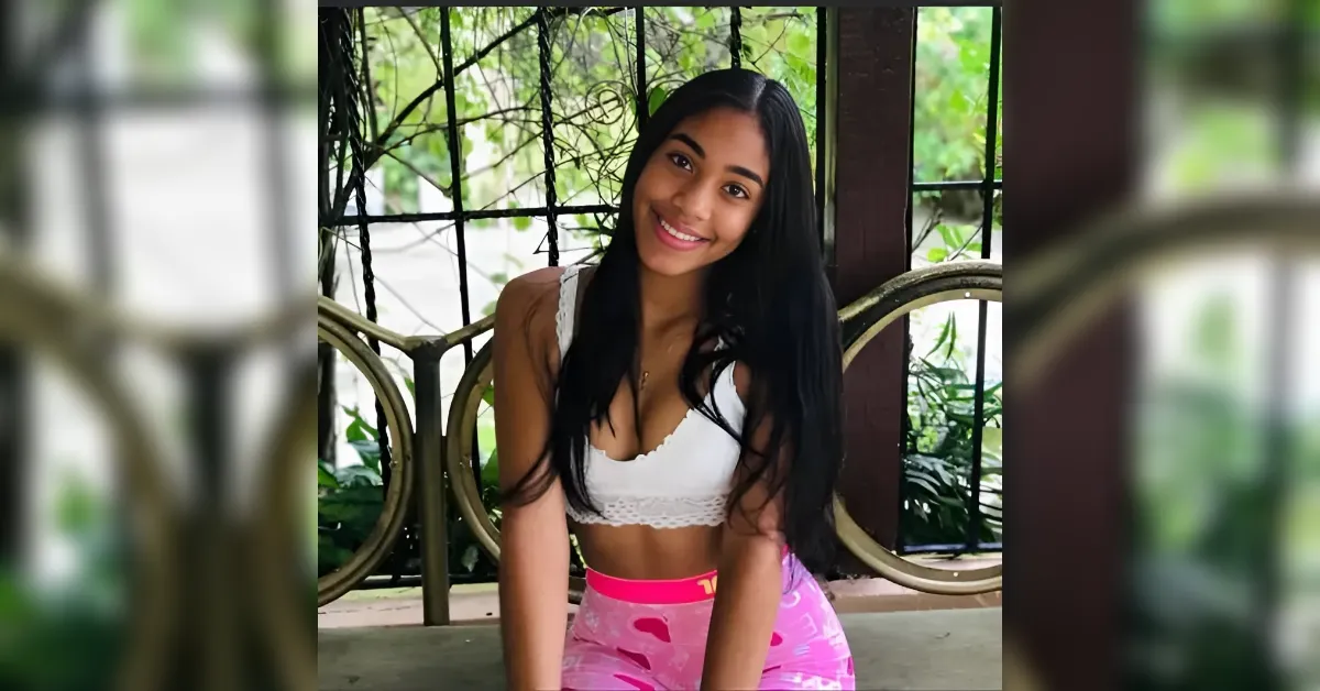 Young woman with long black hair smiling at the camera, wearing a white lace bralette and pink camouflage leggings, leaning on a railing outdoors with greenery behind.