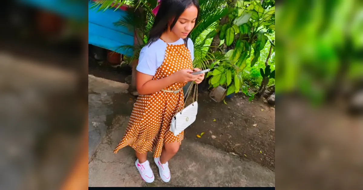 Girl in an orange polka-dot dress and white shirt stands on a path, looking at her smartphone with a white purse hanging from her shoulder.