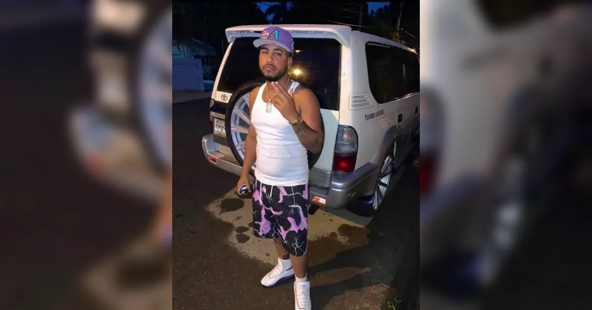 Man in a white tank top, purple cap, and pink camo shorts poses in front of a white SUV at night, flashing a peace sign.