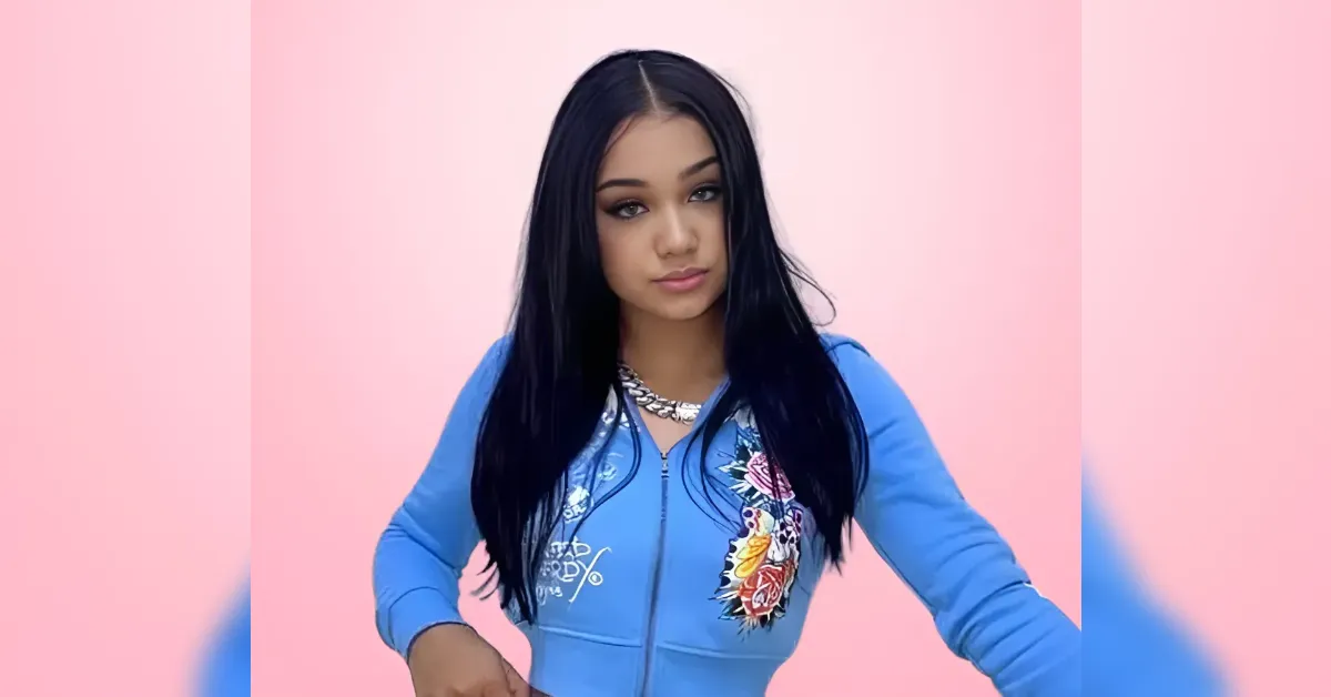 Young woman with long black hair wearing a blue embroidered jacket against a pink backdrop.