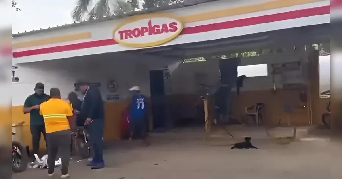 Tropigas storefront with a yellow–red stripe sign; group of people gathered outside, some examining items on a bike or table; a dark dog lying on the ground nearby.