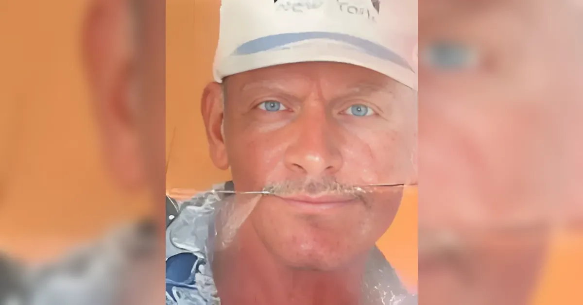 Close-up of a man with blue eyes wearing a white cap, a thin branch held horizontally between his lips against an orange background.