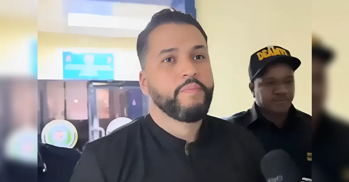 A man with a beard being interviewed indoors, a security guard stands in the background wearing a black cap.