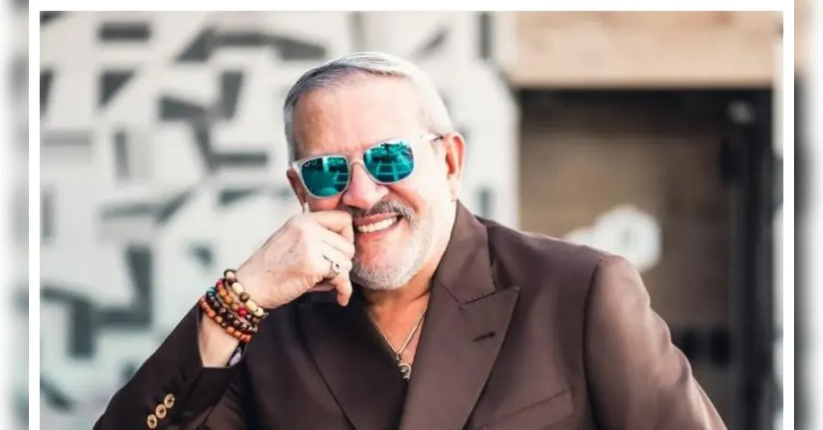 Smiling middle-aged man with gray hair and beard in a brown blazer, wearing blue reflective sunglasses and beaded bracelets, resting his chin on his fist outdoors.