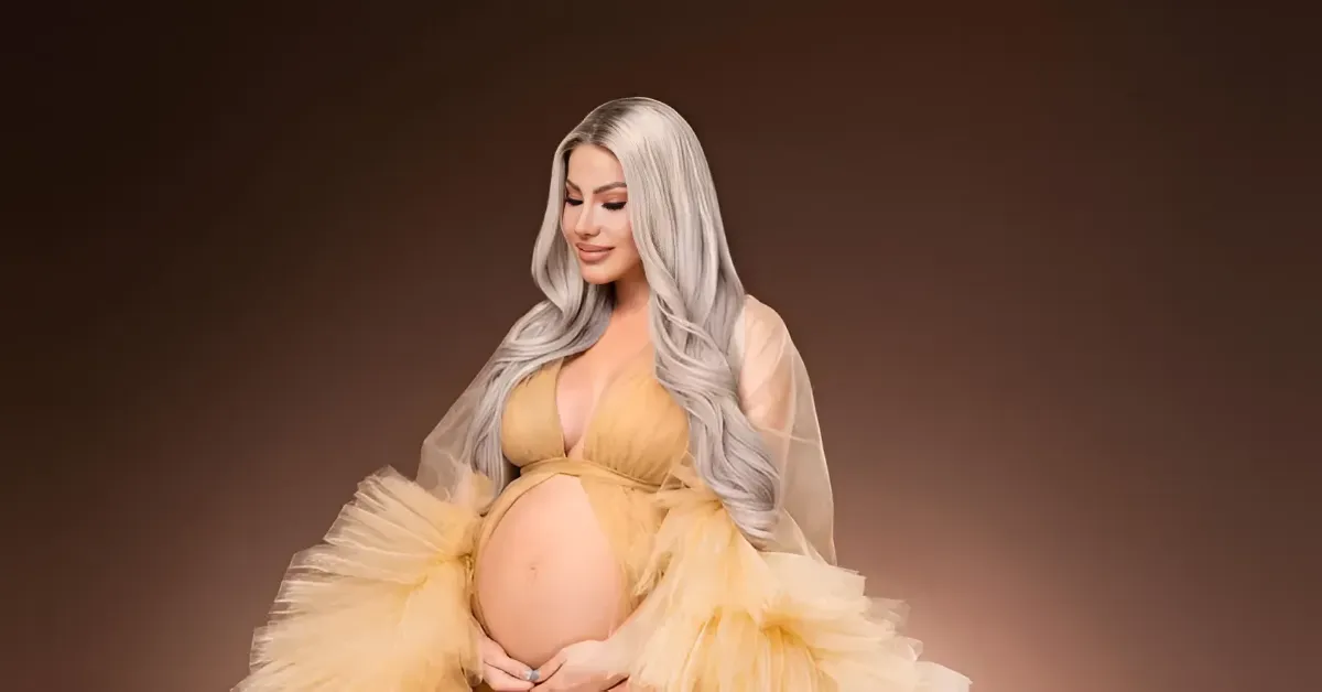 Pregnant woman in a pale yellow gown cradling her baby bump against a warm brown studio backdrop.