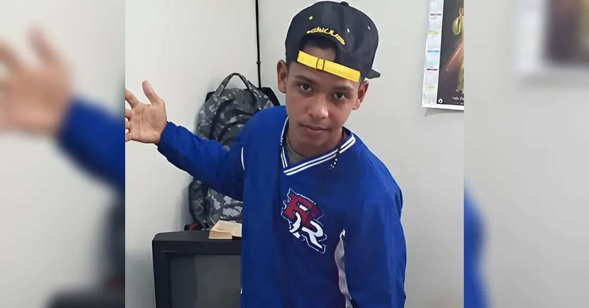 Boy in a blue jacket and a backward black cap with a yellow brim, arm outstretched in a room with a backpack in the background.