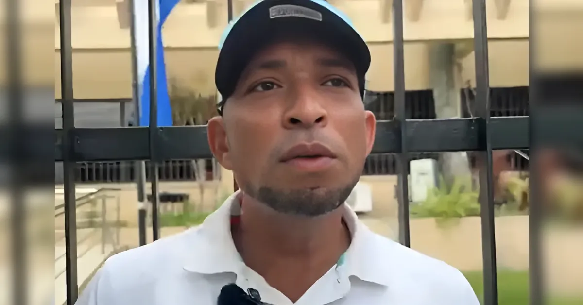 Man wearing a black cap and white shirt speaks to the camera in front of a metal gate outdoors.