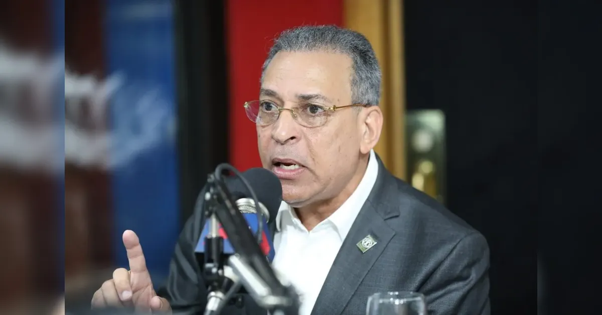 Man in a dark suit and glasses speaks into a microphone during a radio interview, gesturing with his index finger.