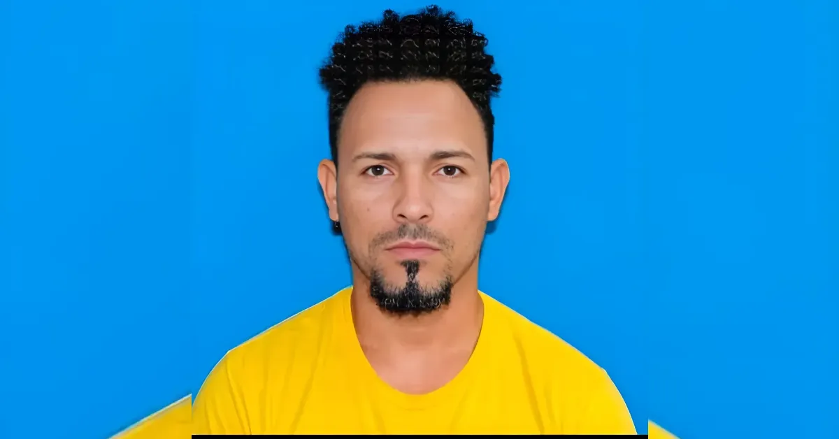 Portrait of a man with short curly black hair and a goatee wearing a yellow shirt against a bright blue background, facing the camera.