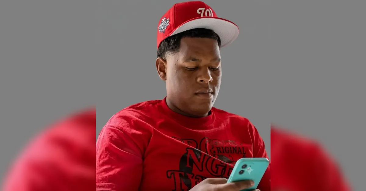 Young man in a red shirt and red cap looking at a teal smartphone outside or in daylight.