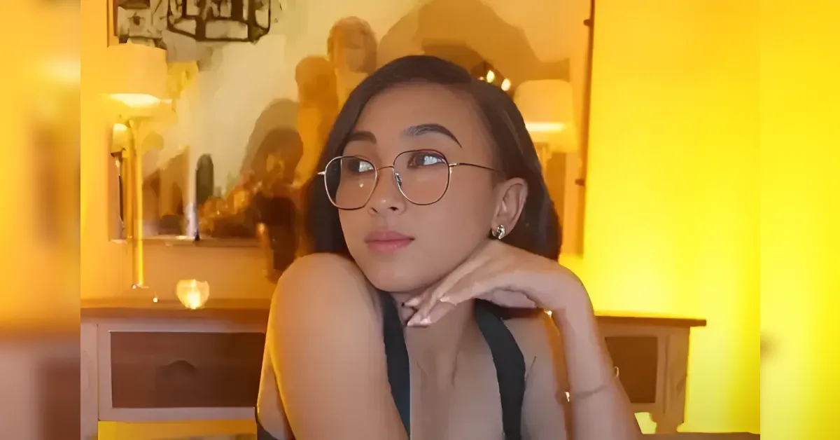 Portrait of a young woman with round glasses resting her chin on her hand in a warm yellow-lit room behind a desk or dresser.