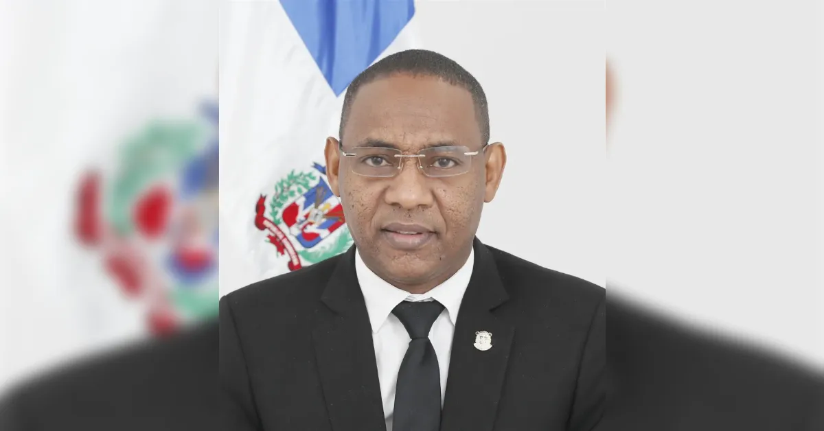 Professional portrait of a man in a black suit, white shirt, and black tie wearing glasses, with a national flag in the background (blurred).