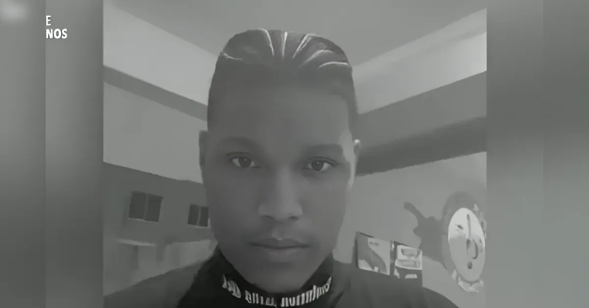 Close-up of a young man with slicked-back hair looking into the camera in a grayscale indoor setting.