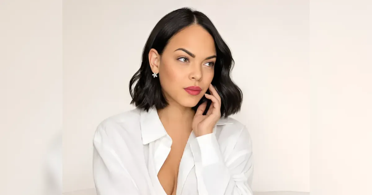 Portrait of a woman with short black hair wearing a white button-down shirt, resting her chin on her hand against a light neutral background.