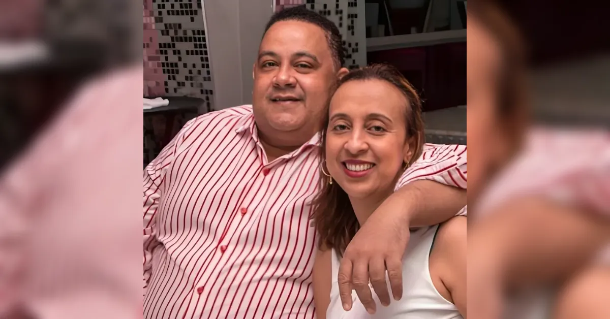 Smiling couple posing closely with the man's arm around the woman, indoors. The man wears a pink striped shirt and the woman a white sleeveless top.