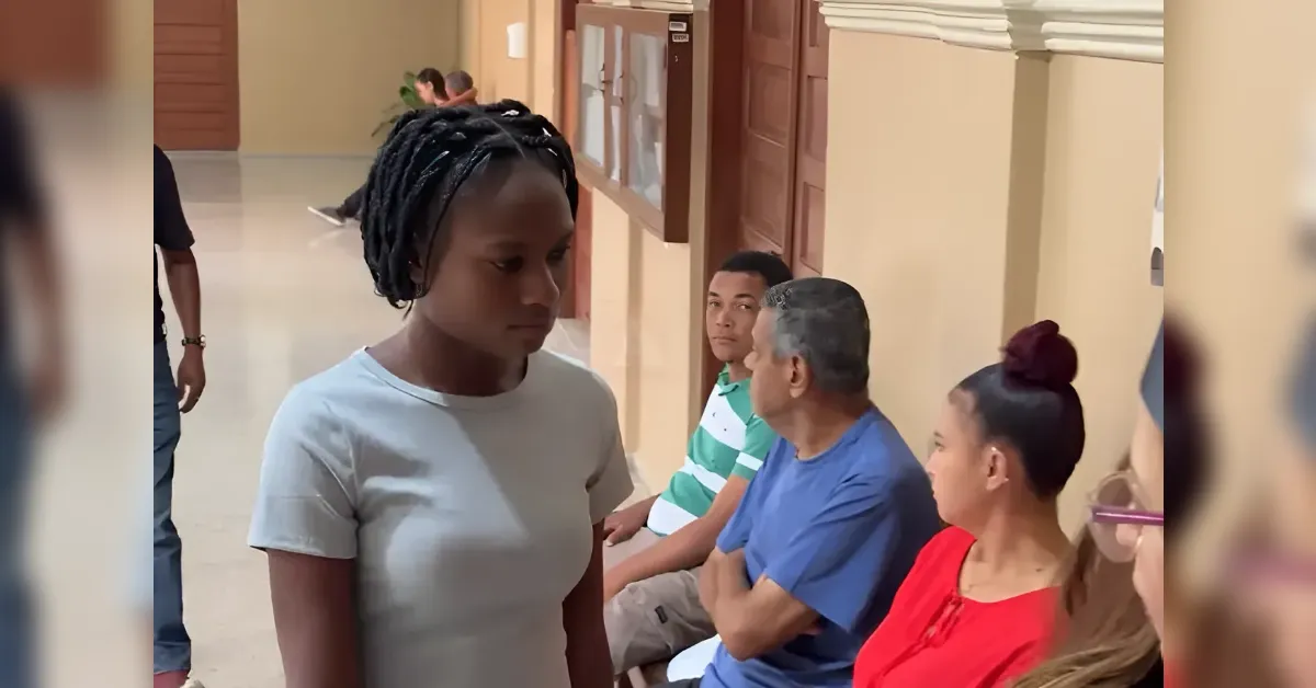 Young woman with braided hair in a light gray shirt walks past seated people in a hallway waiting area.
