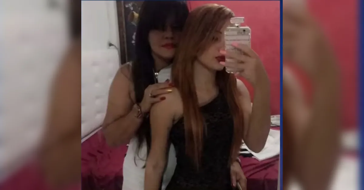 Two women posing for a mirror selfie; one with red hair in front, the other with dark hair behind, in a pink bedroom.",