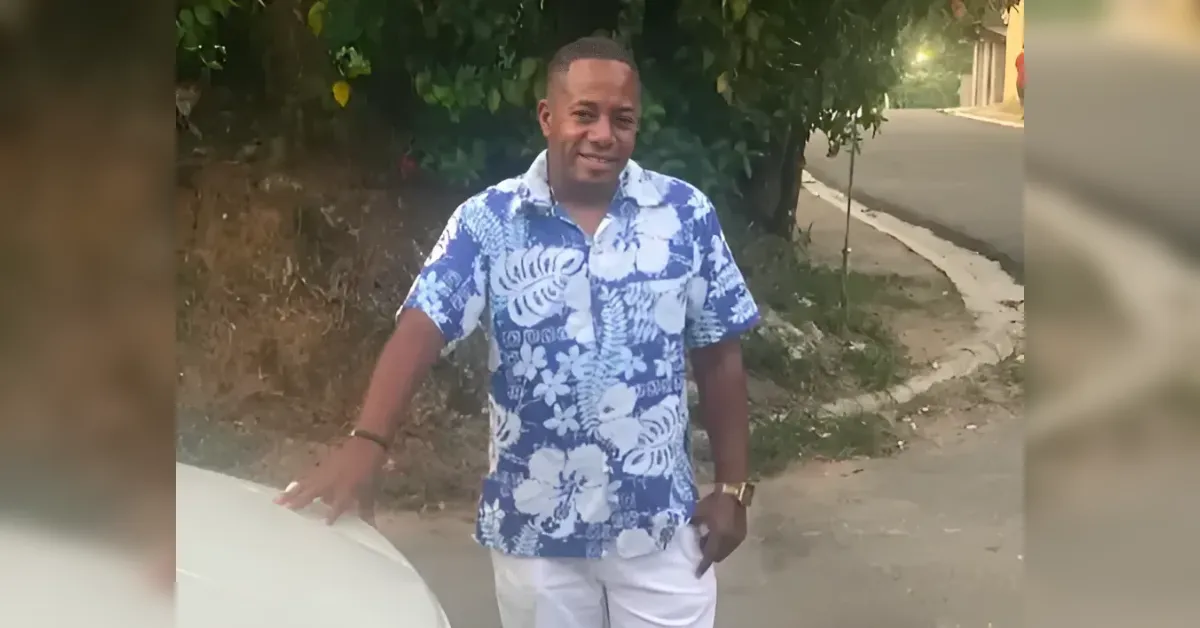 Man in a blue and white tropical shirt standing outdoors next to a car, smiling at the camera on a residential street.