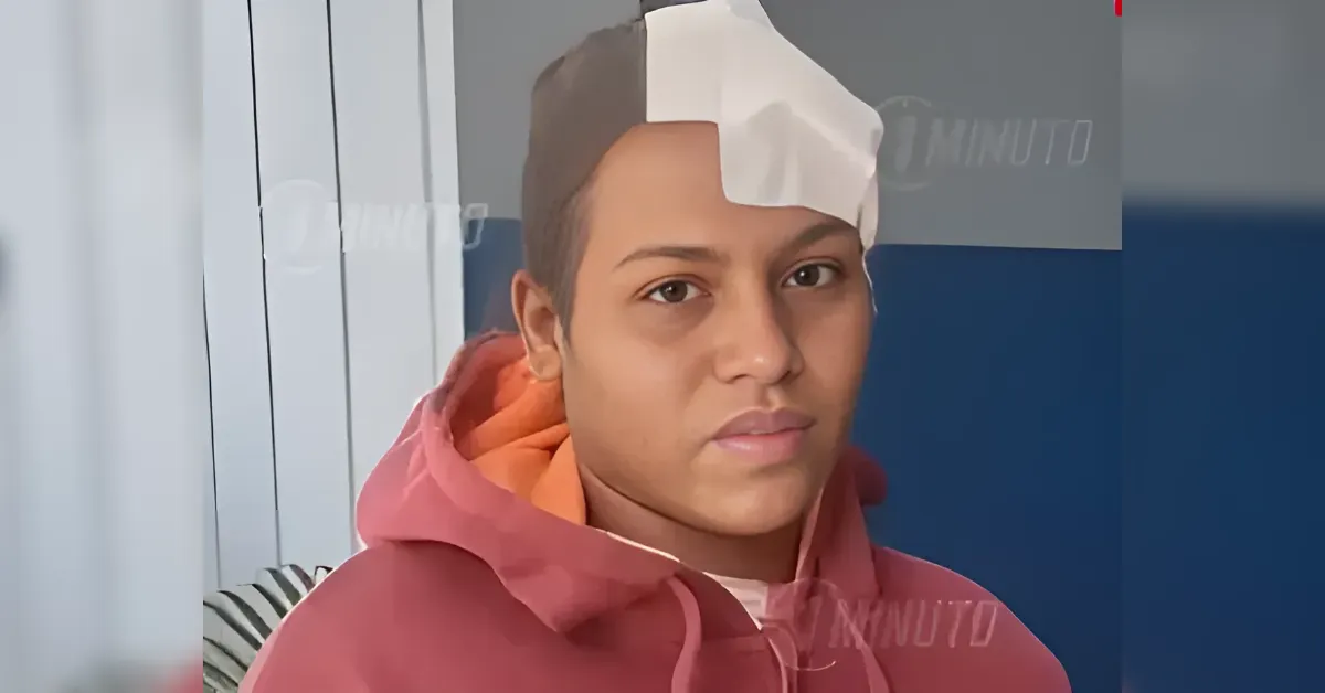 Young man with a white bandage on his head, wearing a pink hoodie, facing the camera indoors.