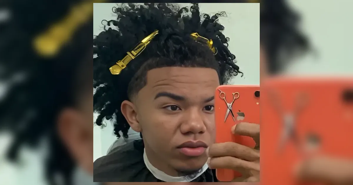 Young man with thick, curly hair and gold hair clips taking a mirror selfie in a barber chair, wearing a black barber cape and a white neck strip, with an orange phone visible in the foreground.