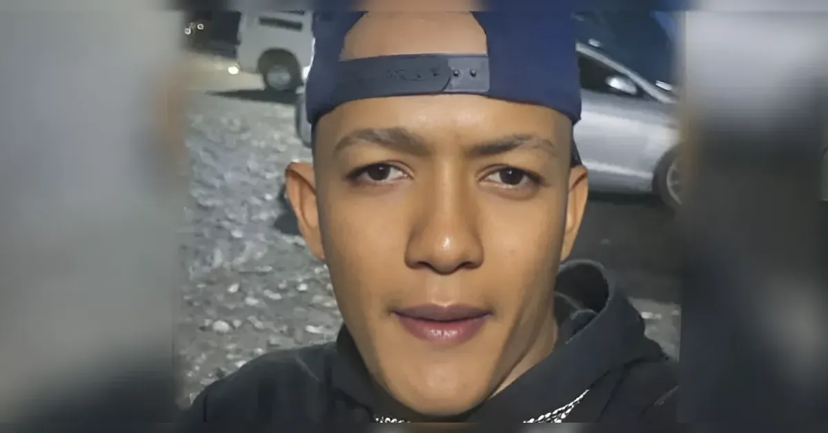 Young man wearing a backward navy baseball cap takes a close-up selfie outdoors, with parked cars in the background.