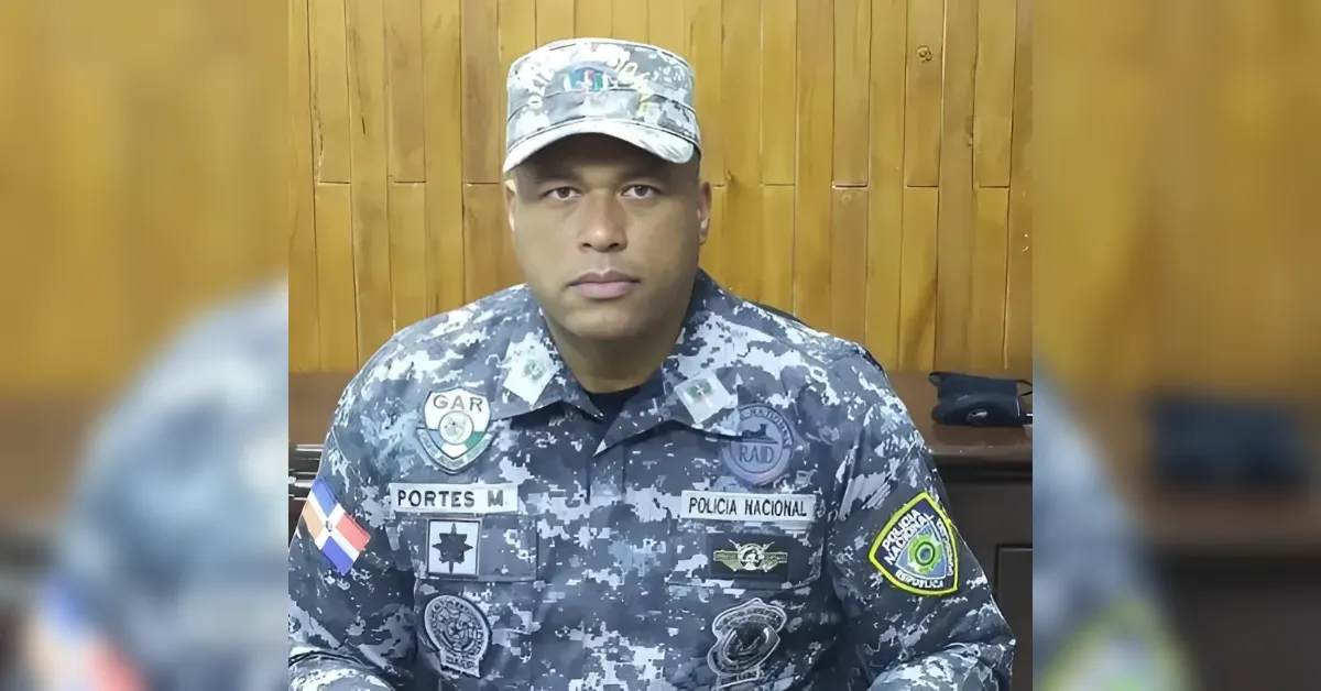 Male police officer in Dominican Republic national police camouflage uniform seated indoors with patches visible on chest and sleeve under a wooden panel backdrop, facing the camera.