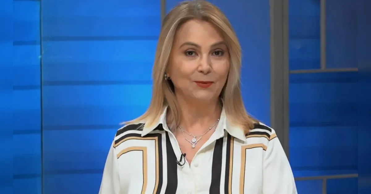 Female news anchor with blonde hair delivering a broadcast in a blue studio backdrop, smiling at the camera.