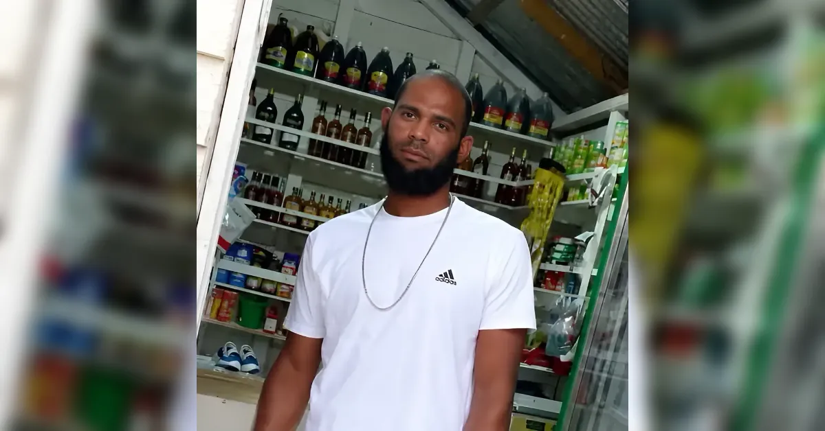 Man with a beard in a white Adidas t-shirt stands inside a shop with shelves of bottles behind him.