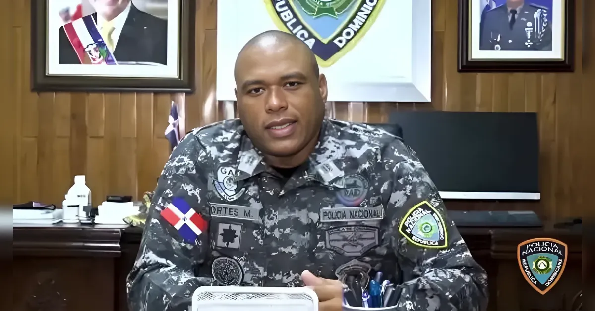 Male police officer in camouflage uniform seated at desk in an office, patches and insignia visible, Dominican Republic flag patch on the chest area, police emblems on wall behind him.