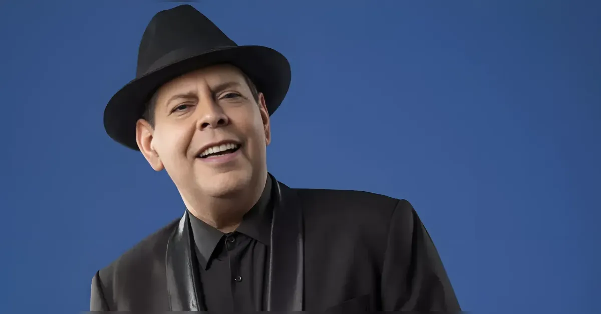 Portrait of a smiling man wearing a black fedora and suit against a solid blue background.