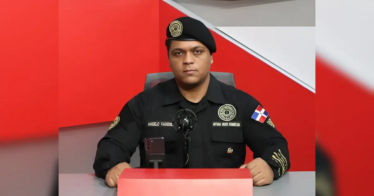 Male security officer in a black uniform with badges, seated at a press desk with a microphone against a red geometric backdrop.
