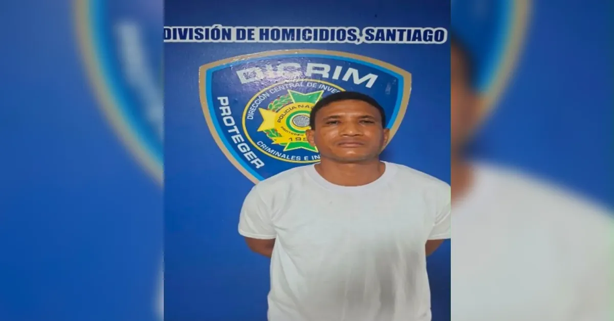 Man in a white T‑shirt stands with hands behind his back in front of a blue police banner with a crest and the words 'División de Homicidios, Santiago'