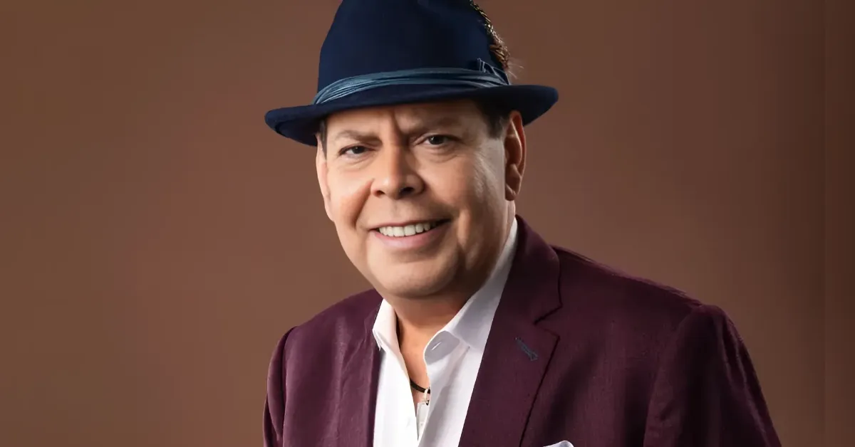 Smiling man in a burgundy blazer and white shirt wearing a navy fedora against a brown backdrop.