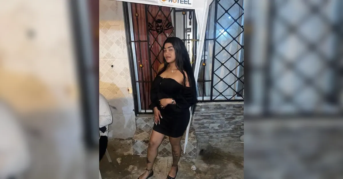 Woman in a black off-the-shoulder dress posing outside a building with a metal gate, looking at the camera.