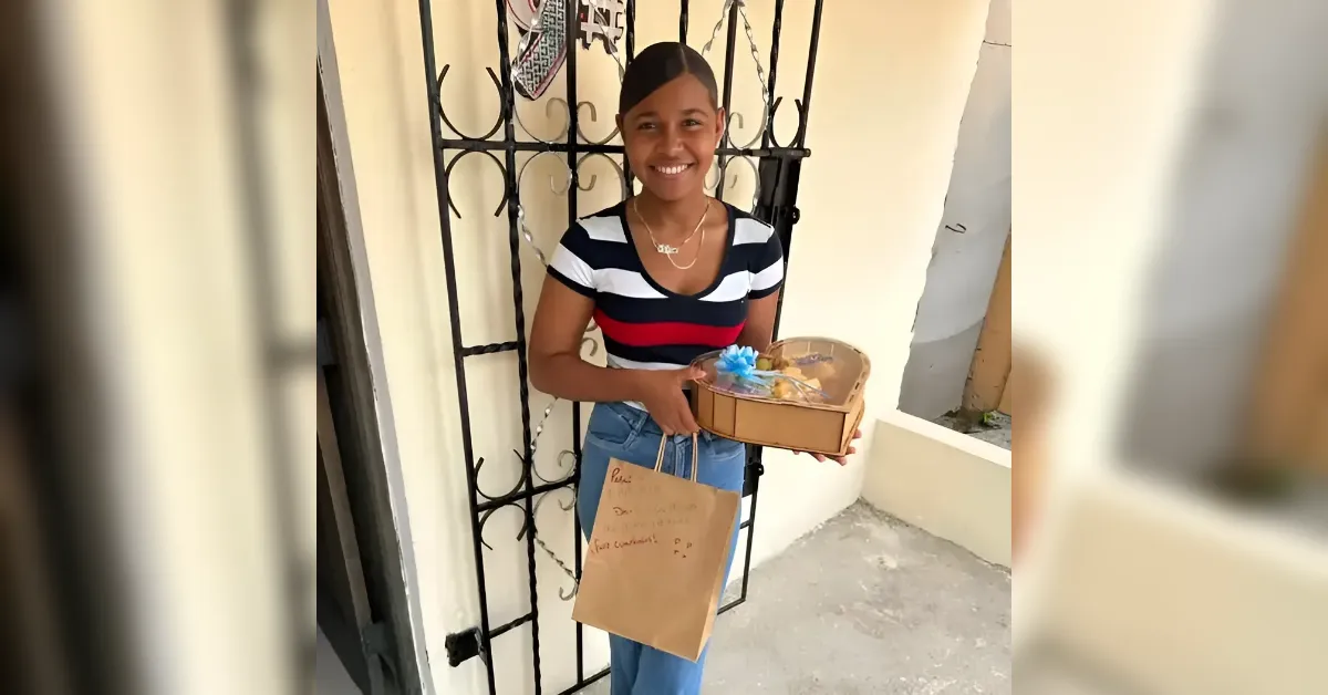 Young woman smiling at a doorway, holding a gift box with a blue bow and a brown paper bag hanging from her arm.