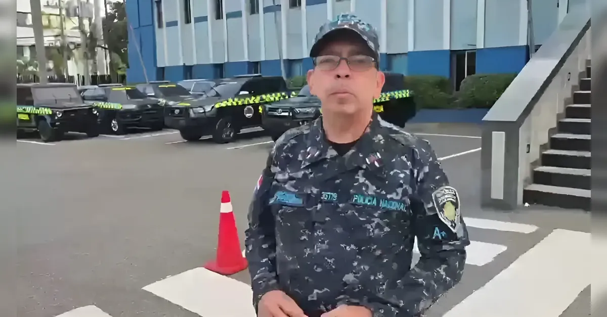 Police officer in camouflage uniform speaking to camera outside a building, with cars taped off in the background by yellow caution tape.