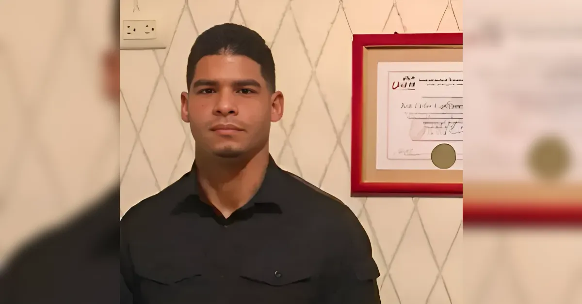 Man in a black shirt standing indoors with a framed document on the wall behind him.