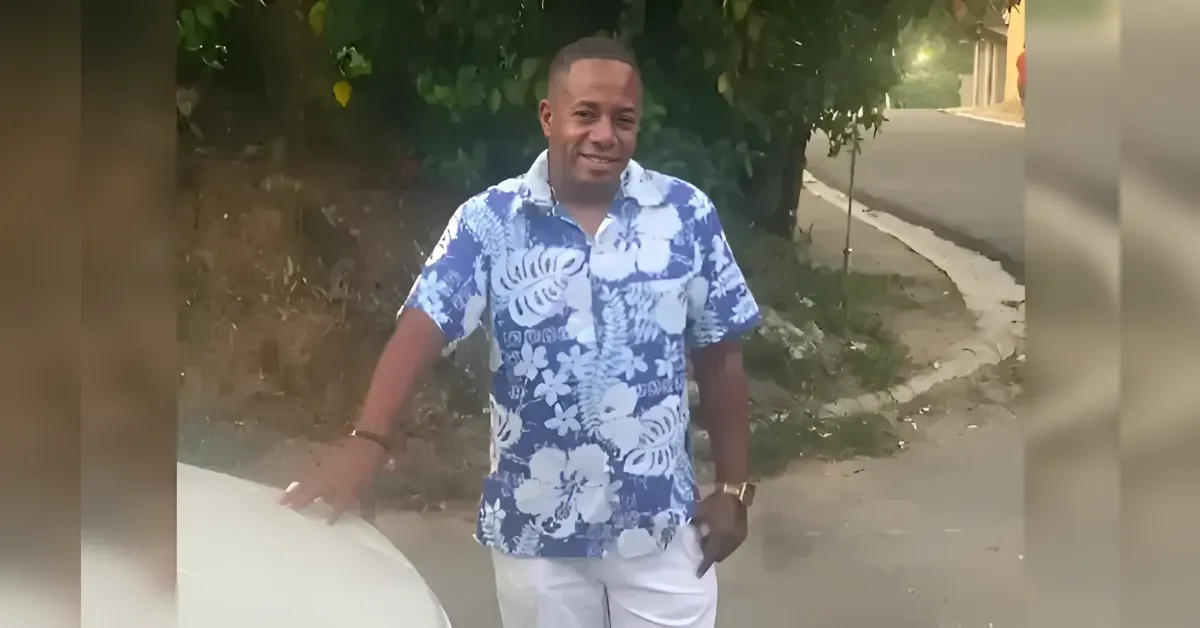 Man in a blue and white floral shirt stands beside a car on a sunny street, smiling at the camera.