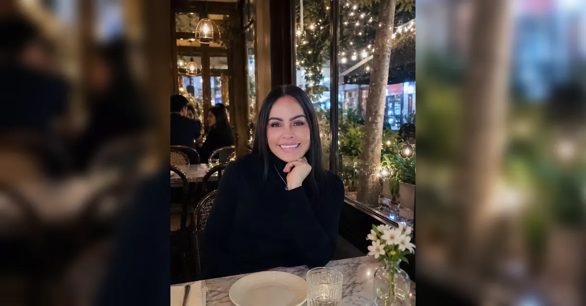 Smiling woman in a black turtleneck sits at a marble-table restaurant by a window with warm string lights and a small vase of flowers.